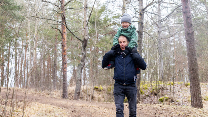 Lapsi istuu aikuisen olkapäillä iloisena metsässä