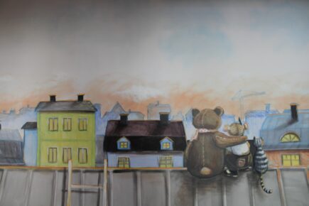 Perheentalon seinällä on seinämaalaus, jossa nalle ja pieni lapsi istuvat vierekkäin vanhan talon katolla taustallaan kaupungin siluetti. Nallen käpälä on lapsen olkapäällä ja tytön vieressä istuu kissa.