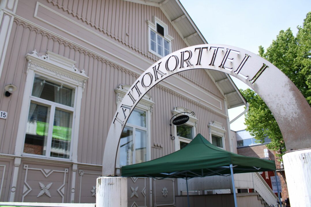 Kuva Perheentalon päädystä pääoven suunnasta kuvattuna. Kuvan etualalla näkyy portti, jonka yläpuolella metallinen kaari tekstillä "Taitokortteli".