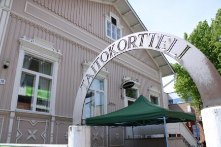 Kuva Perheentalon päädystä pääoven suunnasta kuvattuna. Kuvan etualalla näkyy portti, jonka yläpuolella metallinen kaari tekstillä "Taitokortteli".