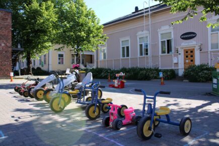 Kuva Perheentalon edestä Taitokorttelin sisäpihalta kesäisin järjestettävältä polkuautopihalta. Kuvassa on erilaisia lasten polkuautoja ja yksi potkumopo rivissä omissa, katuliiduilla piirretyissä parkkiruuduissaan. Taustalla näkyy kartioiden ja katuliiduilla piirrettyjen viivojen avulla tehty ajorata.