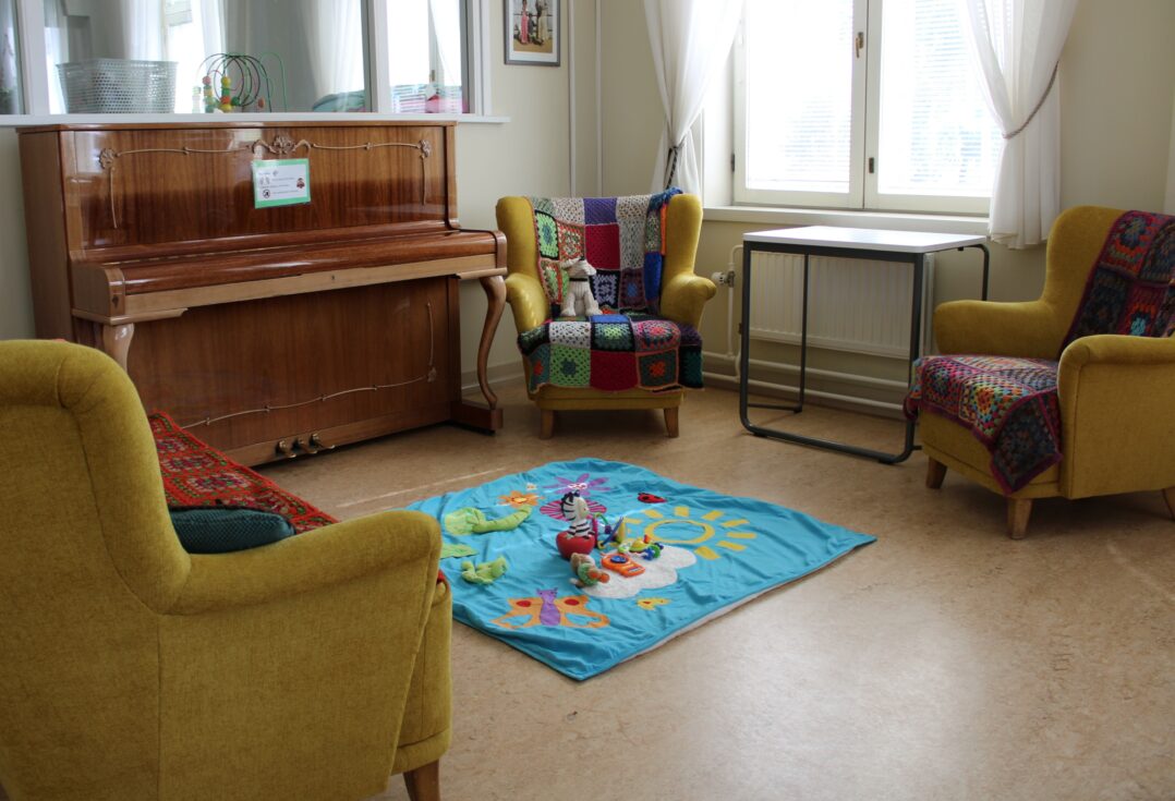 Kuva Perheentalon tuvasta, jossa kaksi nojatuolia ja sohva on aseteltu vastapäätä toisiaan. Piano on tuolien ja sohvan välissä ja keskellä on vauvojen leikkimatto, jonka päällä on leluja.