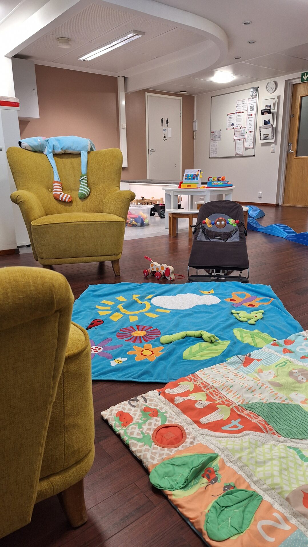 Kuva Perheentalon oleskelu- ja leikkitilasta. Etualalla sohva ja nojatuoli sekä kaksi vauvan peittoa levitettynä lattialle. Taustalla näkyy vauvan sitteri ja leluja.
