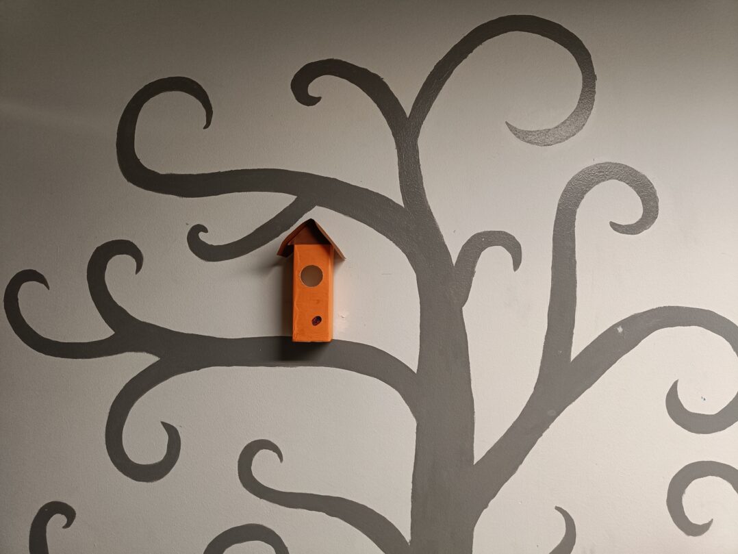 Seinään maalattu lehden puu, sekä pahvinen linnunpönttö yhdellä oksalla