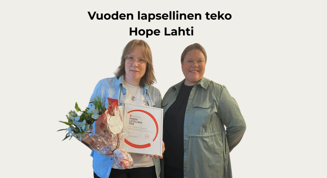 Kuvassa on kaksi hymyilevää naista, joista vasemmanpuolimmainen pitelee käsissään kukkakimppua ja kunniakirjaa. 