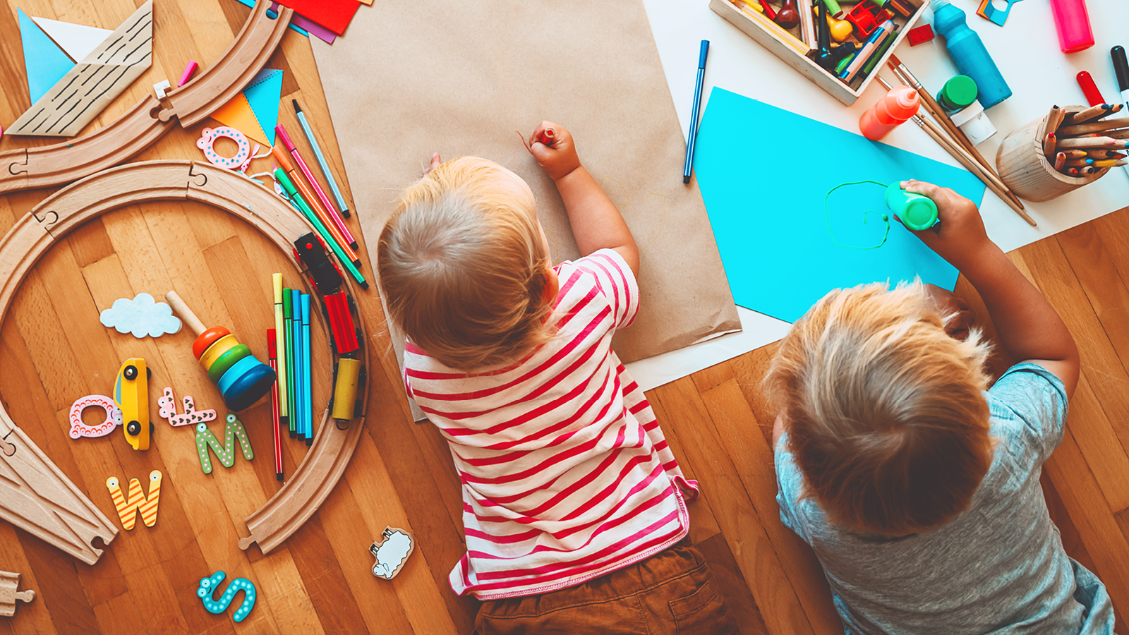 Kaksi lasta piirtämässä ja maalaamassa lattialla maaten värikkäille papereille.