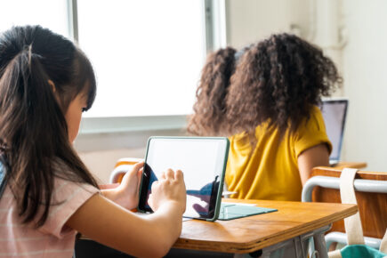 Elementary school girls using digital device in school classroom, digital native, technology, learning, touchscreen. Female students in class from behind.