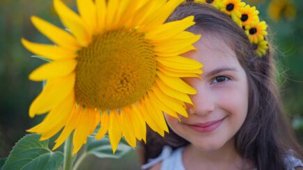 kukkaseppeleinen tyttö hymyilee ison auringonkukan takaa