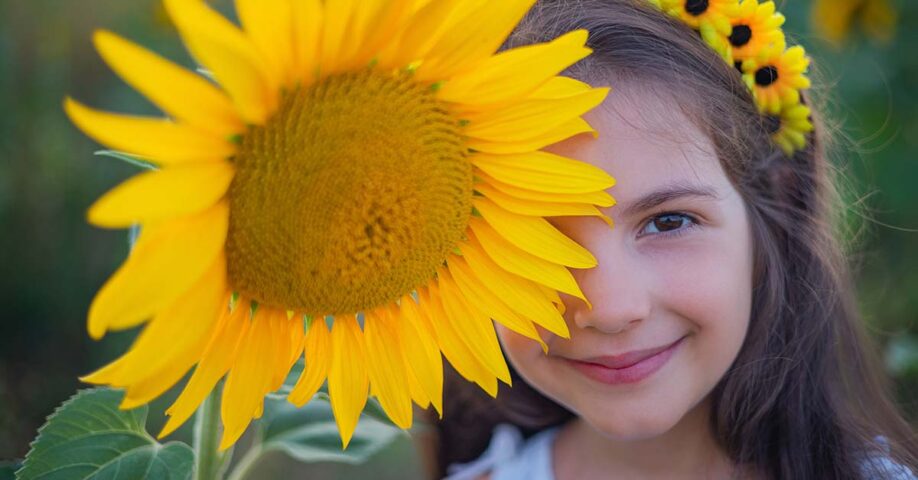 kukkaseppeleinen tyttö hymyilee ison auringonkukan takaa