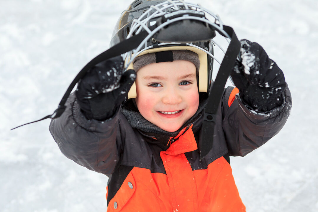 Kuvassa iloinen lapsi kurkistaa jääkiekkokypärän visiirin alta lumisessa maisemassa.