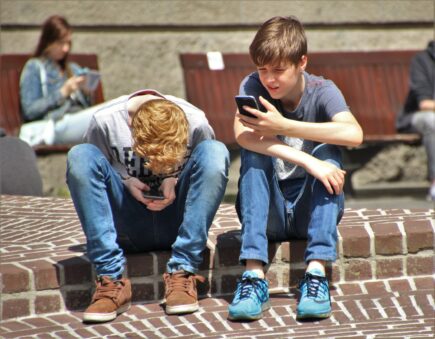 Kuvituskuva, jossa kaksi lasta istuu katukivetyksellä vierekkäin ja katsovat puhelimia käsissään.