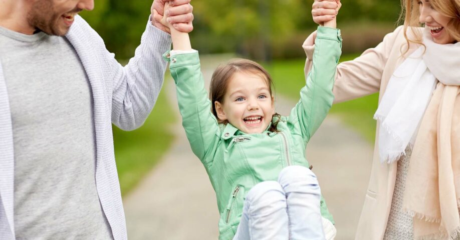 vanhemmat nostaa nauravaa vihreä takkista tyttöä käsistä ylös ulkona