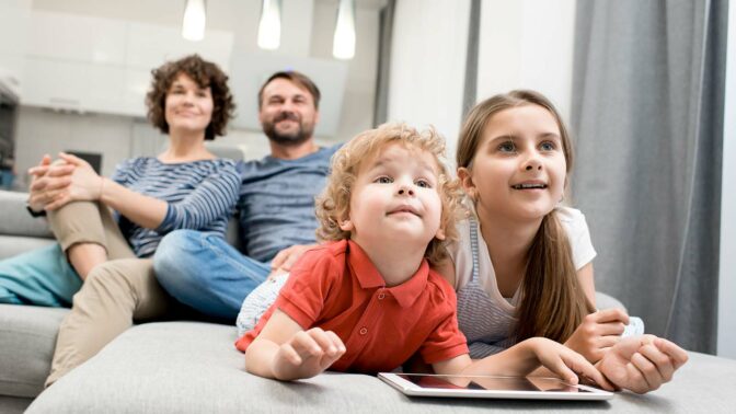 Perhe katselee televisiota sohvalla, lapset edessä ja vanhemmat taka-alalla.