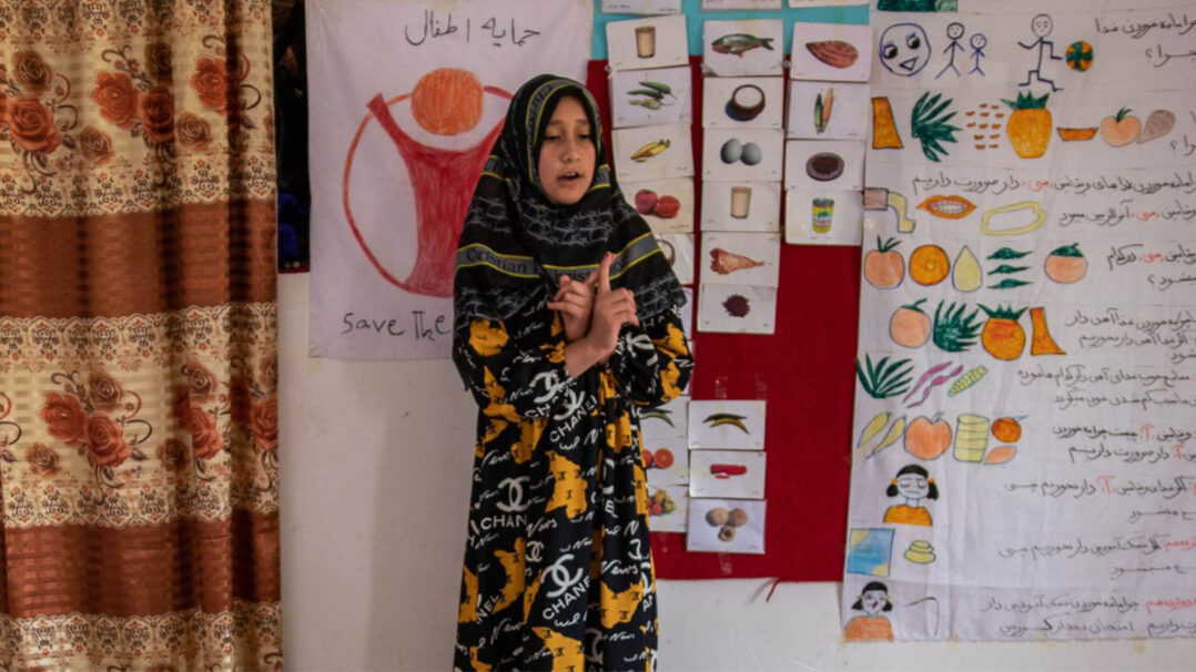 Nuori tyttö puhuu koululuokan edessä taustallaan erilaisia kuvallisia opetusmateriaaleja ja piirretty Pelastakaa Lapset -logo