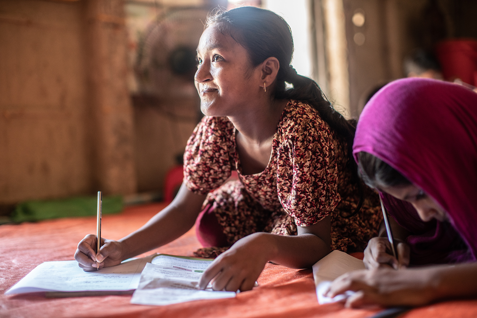 Kaksi tyttöä istuvat lattialla ja kirjoittavat kirjaansa Bangladeshissa.
