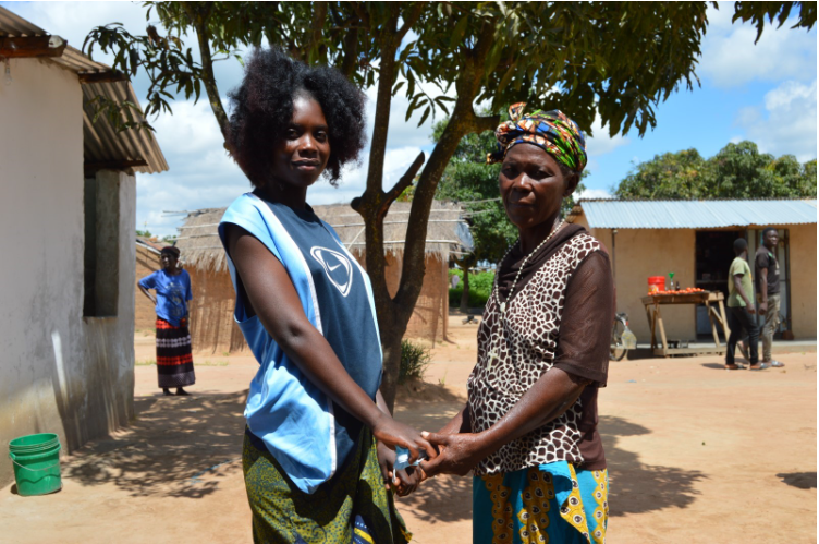Nainen pitää äitiään kädestä ulkona talon edessä Sambiassa.