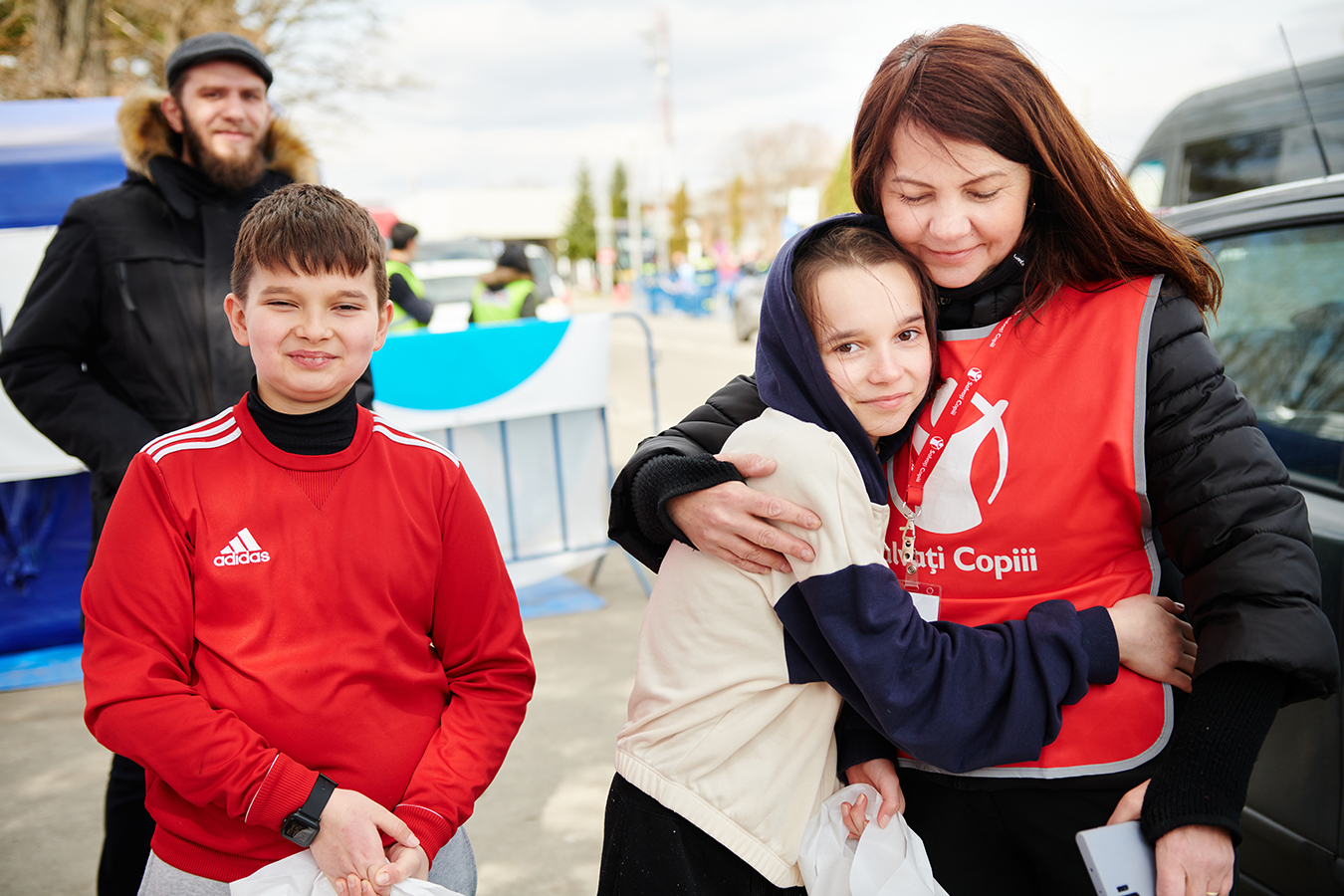 Nainen, jolla on Pelastakaa Lasten liivi halaa hymyilevää tyttöä. Vieressä hymyilevä poika ja taustalla mies. Ukrainan ja Romanian rajalla.