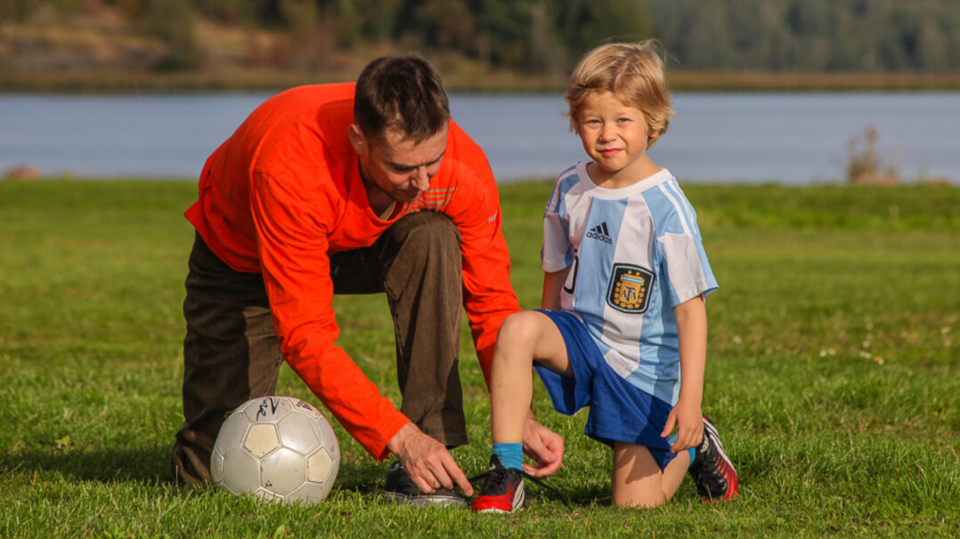 Mies ja poika kesäisellä nurmikolla jalkapallon kanssa ja mies solmii pojan kengännauhoja.