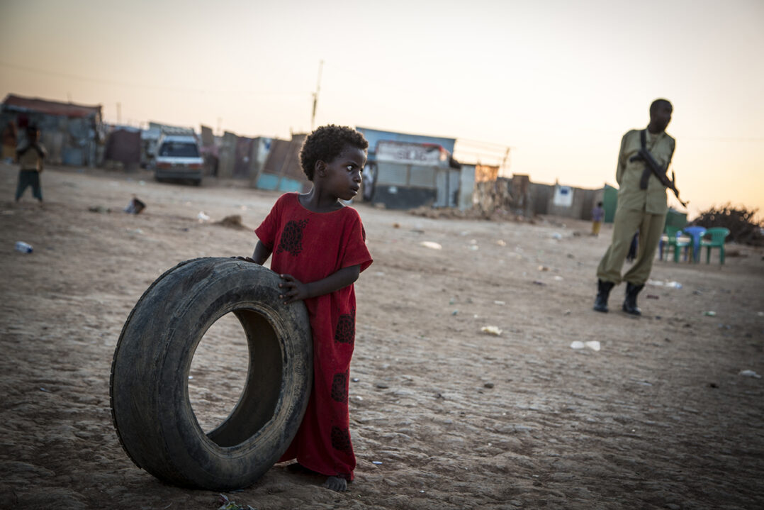 Lapsi pitelee autonrengasta käsissään ja katsoo sivulla. Taustalla näkyy pakolaisleiri ja mies aseen kanssa.