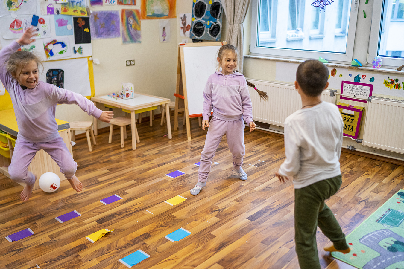 Kolme lasta leikkii lapsiystävällisessä tilassa. Yksi lapsista on hypännyt ilmaan ja yksi seisoo yhdellä jalalla. Laittialla on värikkäitä lappuja.