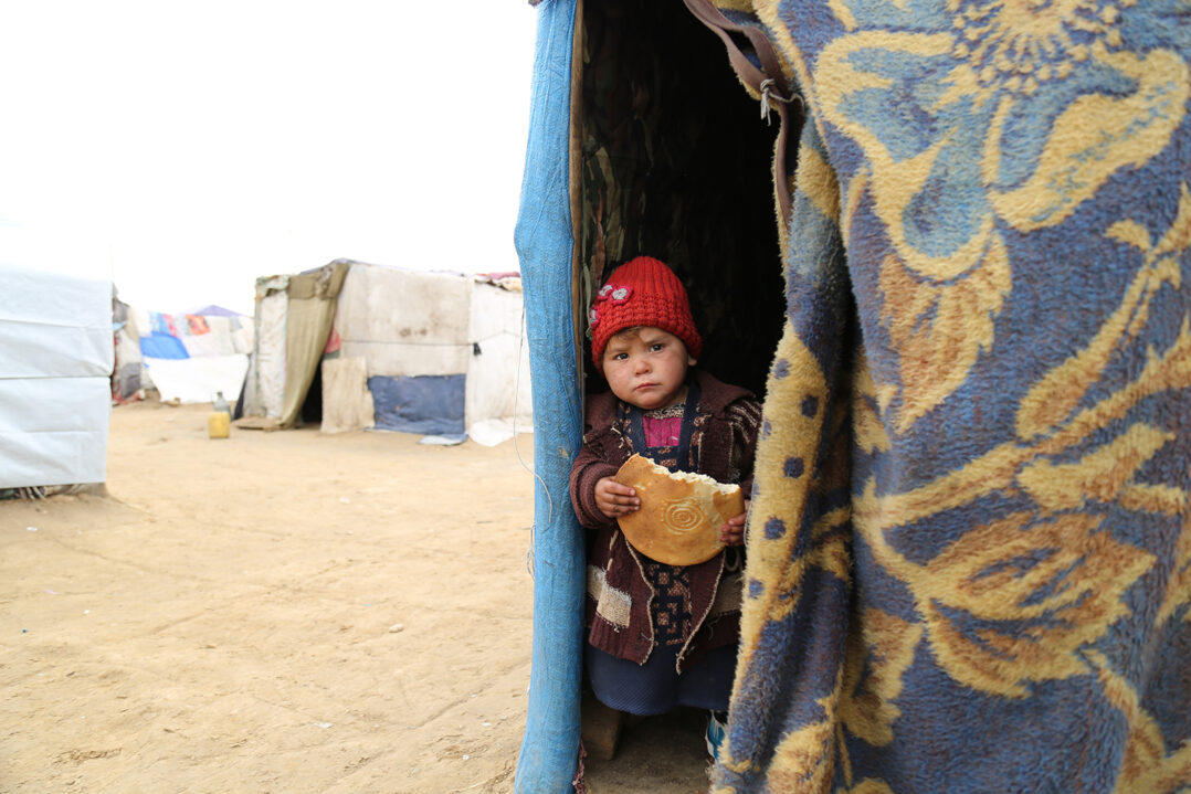 Pieni lapsi kurkistaa ulos teltasta pakolaisleirillä. Hänellä on leipä kädessä.
