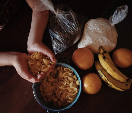 Kulho, jossa on pastaa ja kulhon yläpuolella on kädet, joissa pastaa. Pöydällä kulhon vieressä appelsiinejä, banaaneja ja riisiä