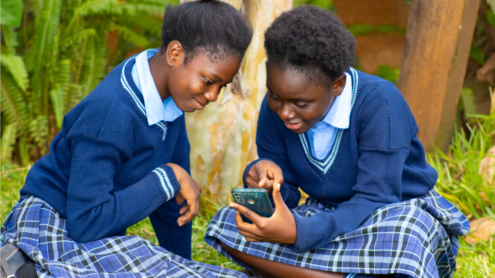 Tytöt istuvat nurmella ja katsovat kännykkää Sambiassa