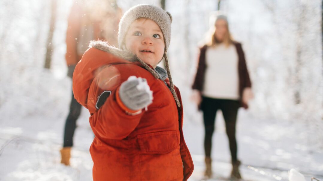 Pieni lapsi talvivaatteissa lunta kädessä, taustalla kaksi aikuista.