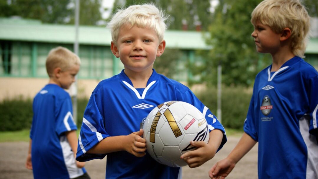 Lapsi sinisessä pelipaidassa jalkapallo kainalossa, taustalla kaksi muuta lasta samanlaisissa paidoissa.