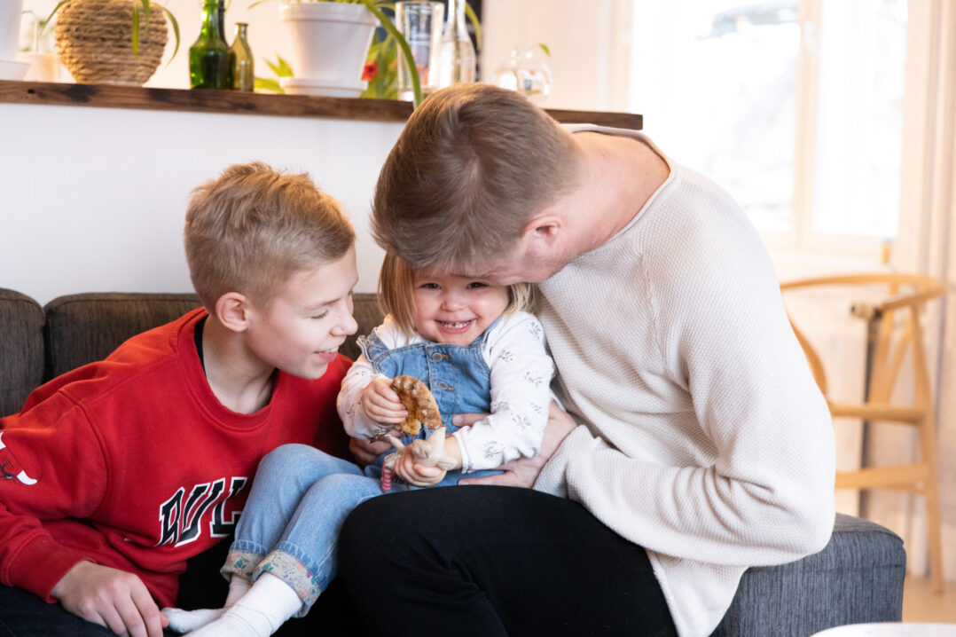 Pieni tyttö nauraa aikuisen sylissä ja syö riisipiirakkaa, vieressä vanhempi poika.