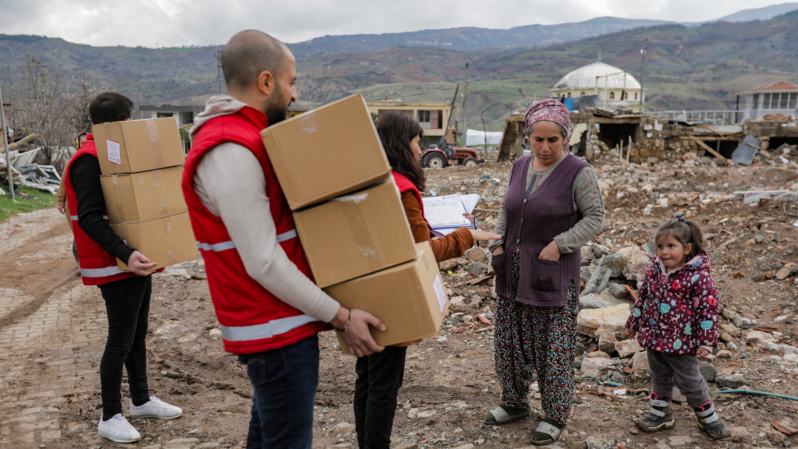 Pelastakaa Lasten työntekijät kantavat pahvilaatikoita, joissa hätäaputarvikkeita. Heidän vierellään on nainen ja lapsi Turkin maanjäristykset ovat tuhonneet taustalla olevaa maisemaa.