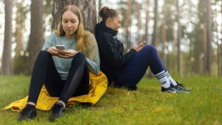 Kaksi nuorta istuu metsässä puun juurella ja käyttää kännyköitään.