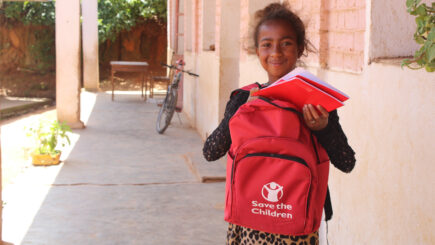 Hymyilevä tyttö seisoo koulun käytävällä Pelastakaa Lapset repun ja koulukirjan kanssa.