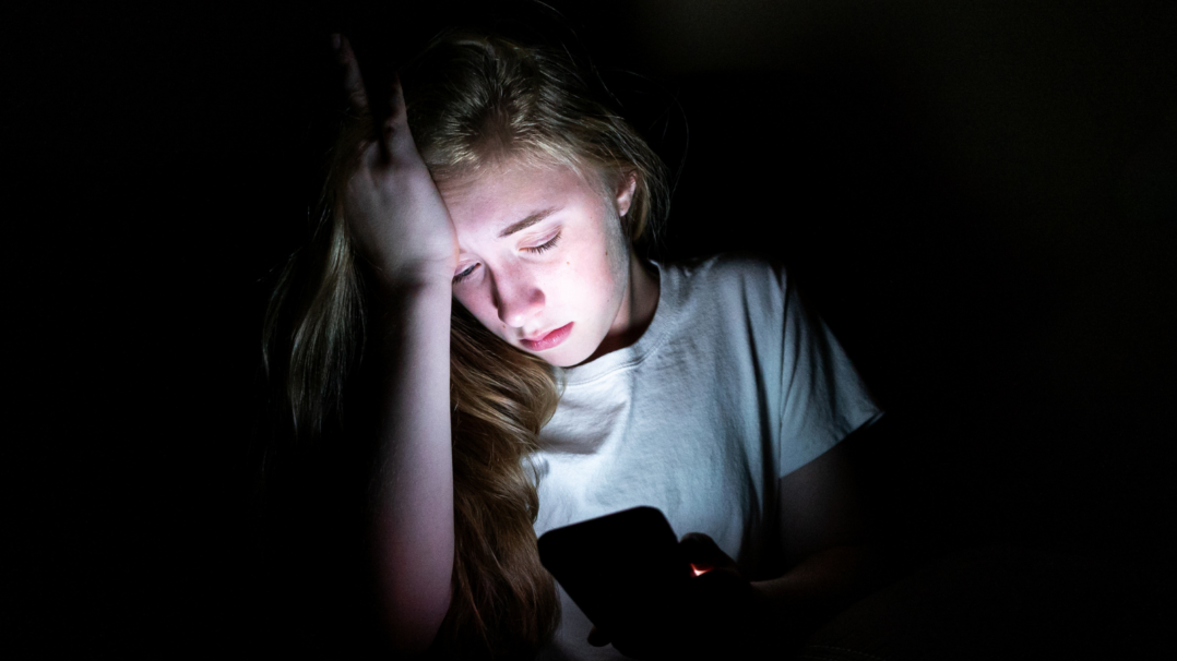 Nuori istuu masentuneena pimeässä matkapuhelimensa kanssa.