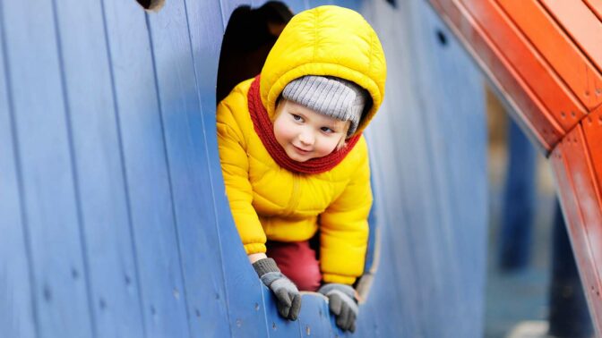 Keltaisessa toppatakissa oleva lapsi kurkistaa leikkipuiston mökin ikkunasta
