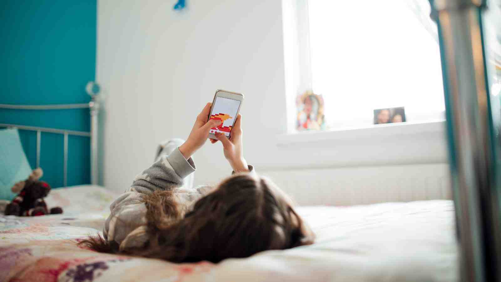 Lapsi ma.koilee selällään sängyllä älypuhelin kädessä