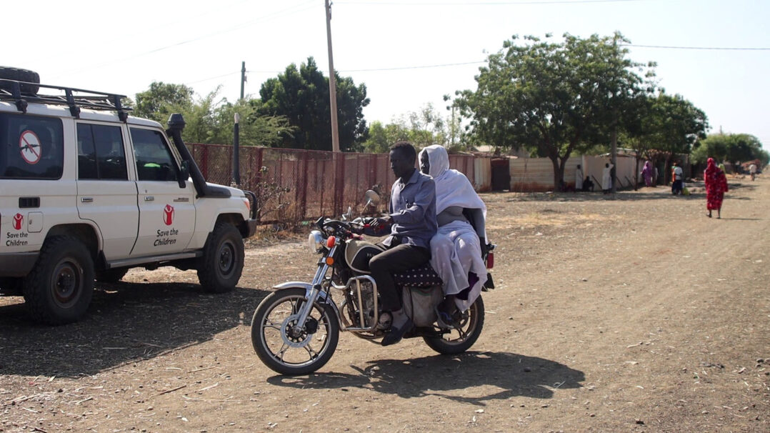 Mies ja nainen ajavat mopolla hiekkatiellä Sudanissa. Taustalla Pelastakaa Lasten jeeppi ja ihmisiä kävelemässä tiellä.