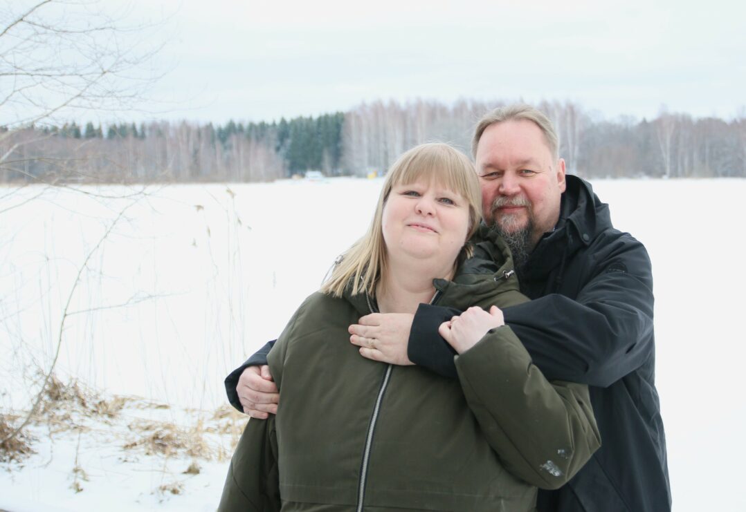 Nainen ja mies seisovat lumisen maiseman edessä. Mies on kietonut kätensä naisen ympärille.