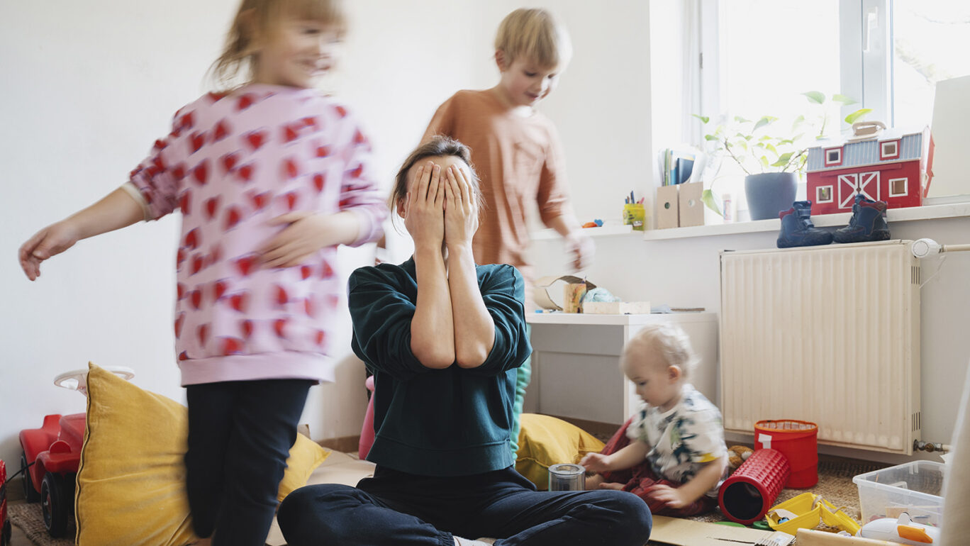 Äiti istuu lattialla kolmen lapsen ympäröimänä ja leikkii piilosta