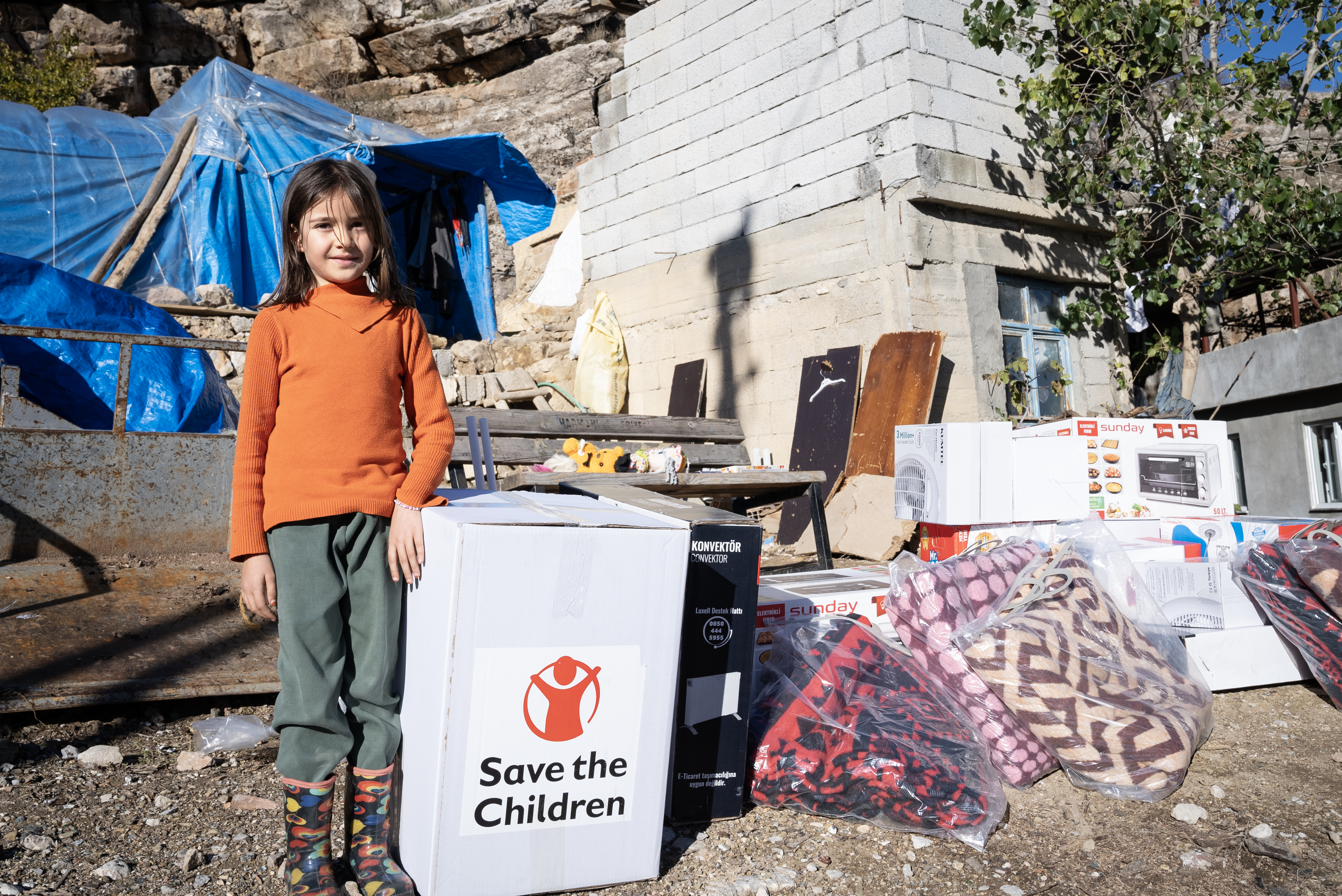 Tyttö seisoo raunioiden keskellä vieressään Save the Children -avustuslaatikko.