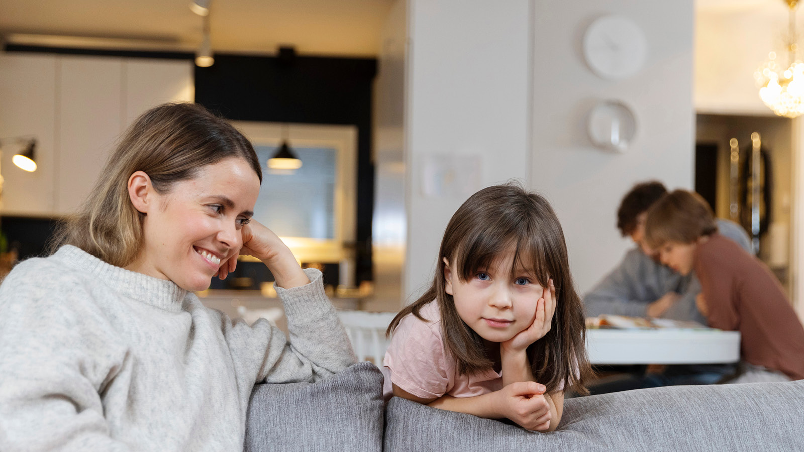 nainen katselee tyttöä ja hymyilee tälle. Tyttö katsoo vakavana suoraan kameraan. Taustalla poika ja mies katsovat yhdessä kirjaa.