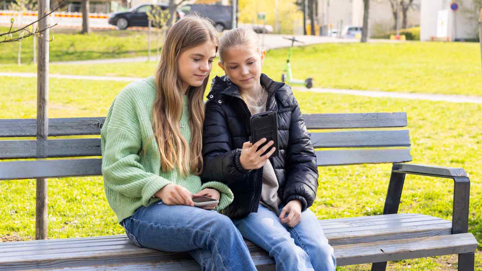Kaksi lasta istuu ulkona penkillä ja kuvaa itseään kännykällä.