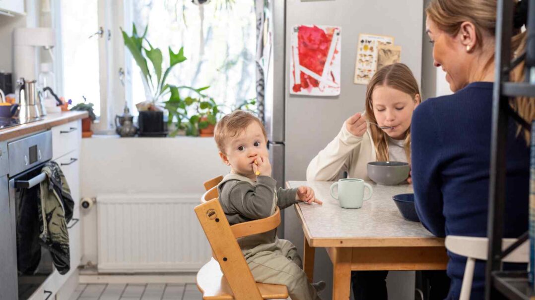 Äiti ja kaksi lasta keittiössä ruokapöydän ääressä. Lapset syövät ja nuorempi katsoo syöttötuolista kohti kameraa.