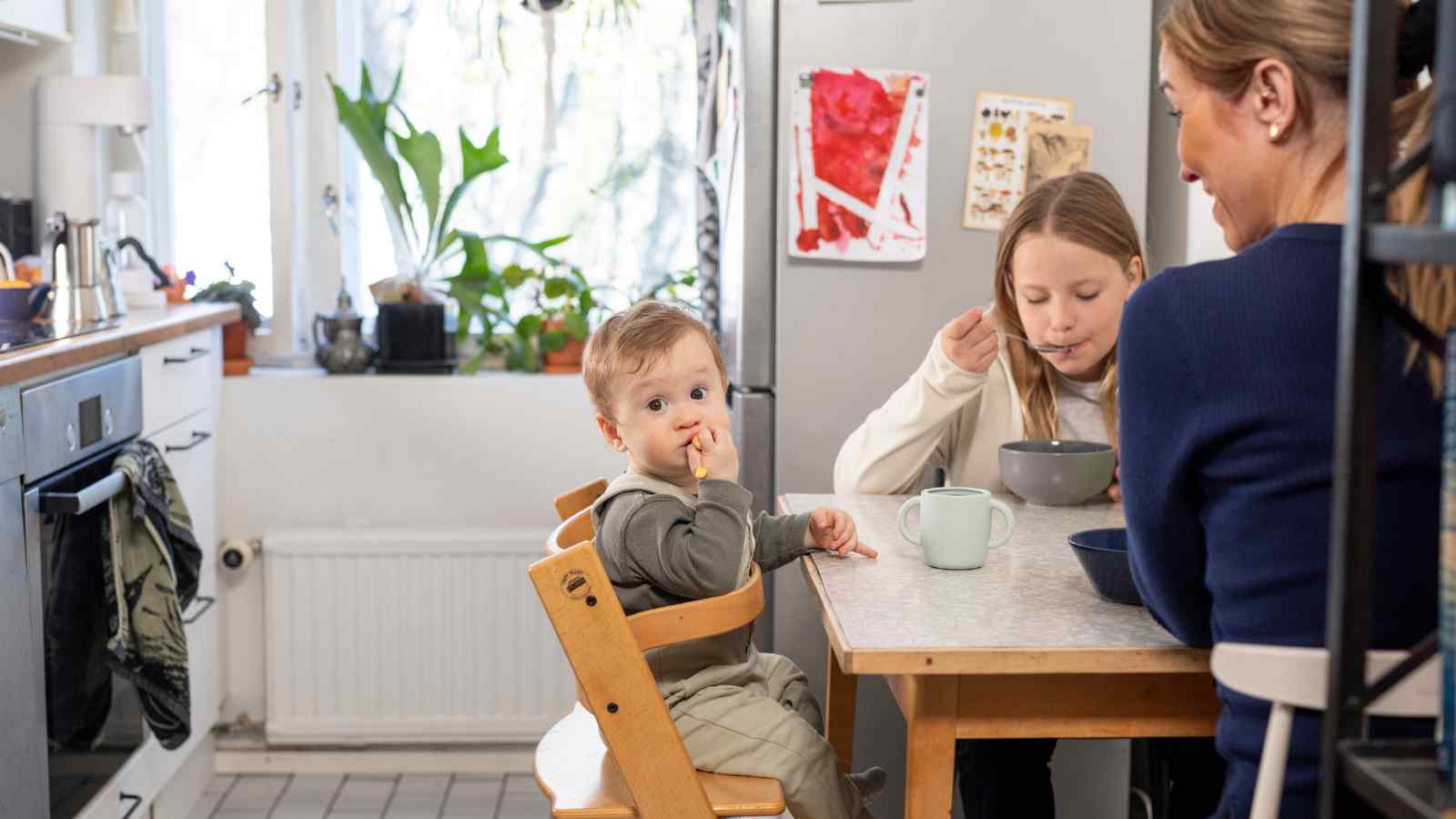 Äiti ja kaksi lasta keittiössä ruokapöydän ääressä. Lapset syövät ja nuorempi katsoo syöttötuolista kohti kameraa.