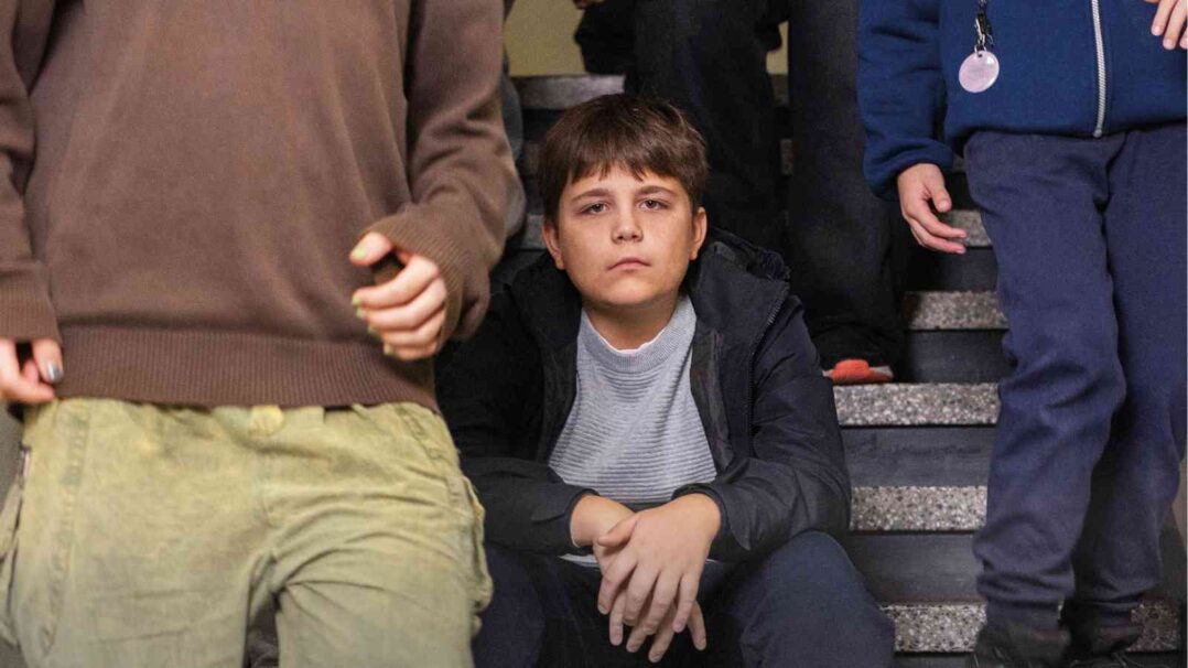 Lapsi istuu portaalla ja katsoo kameraan muiden kävellessä ohi.