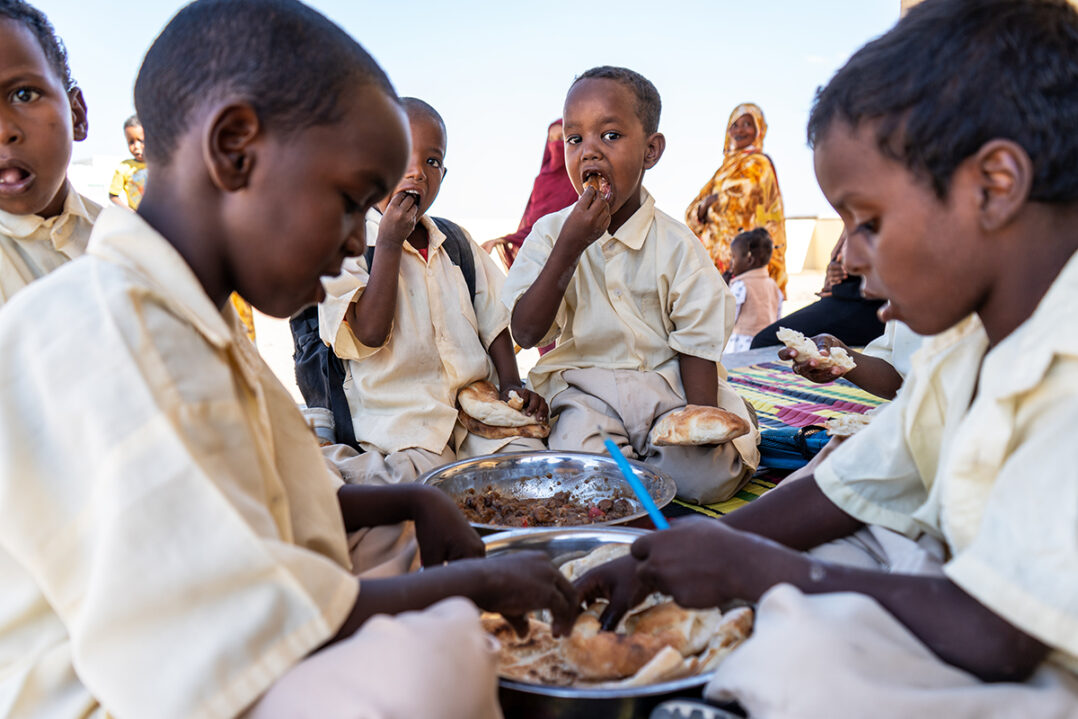 Koululaiset ruokailemassa yhteisen aterian äärellä vaaleat vaatteet päällään.