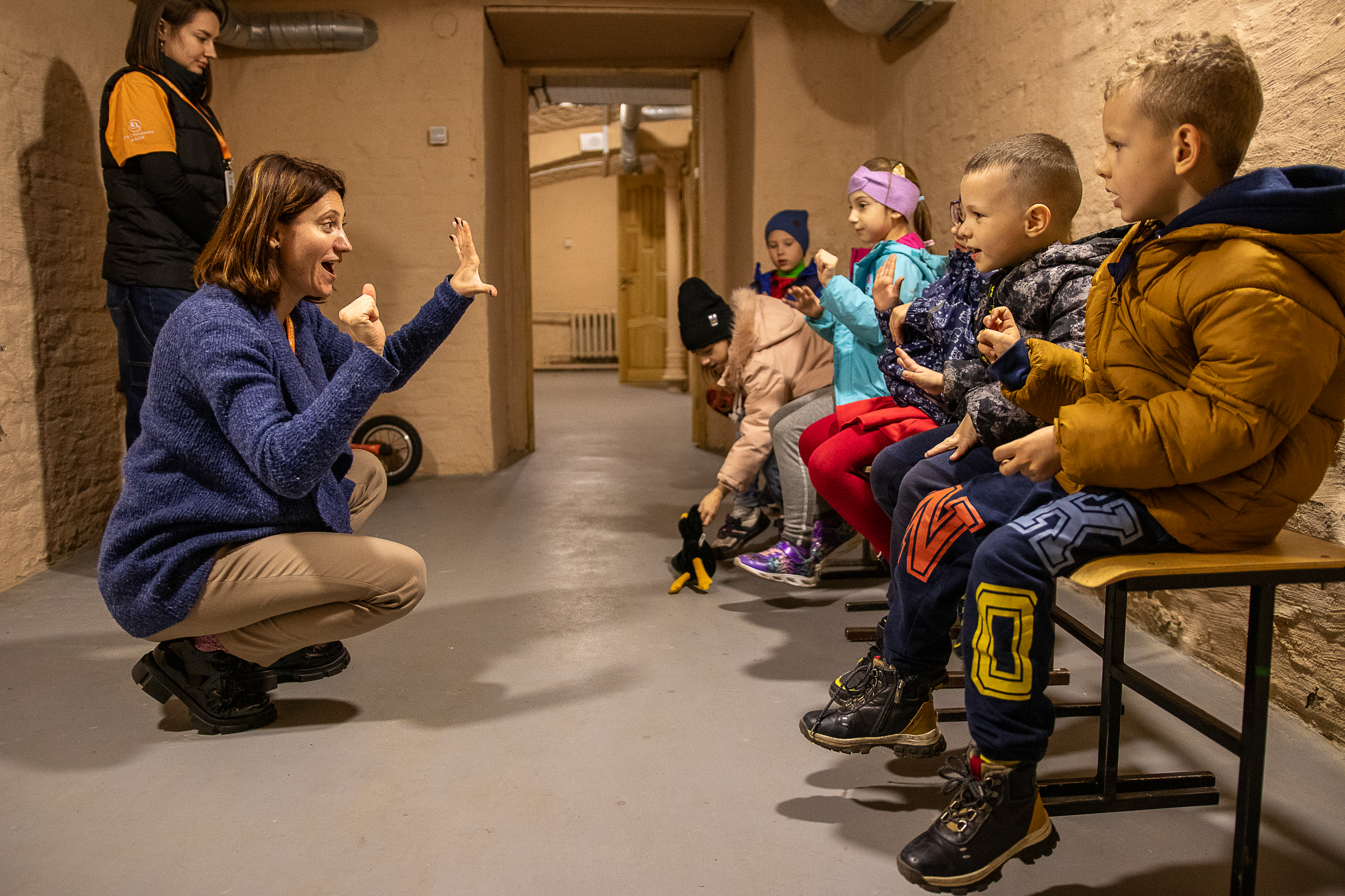 Lapsia istuu väestönsuojassa takit päällään. Vastapäätä aikuinen opettaa tai kertoo lapsille jotain.