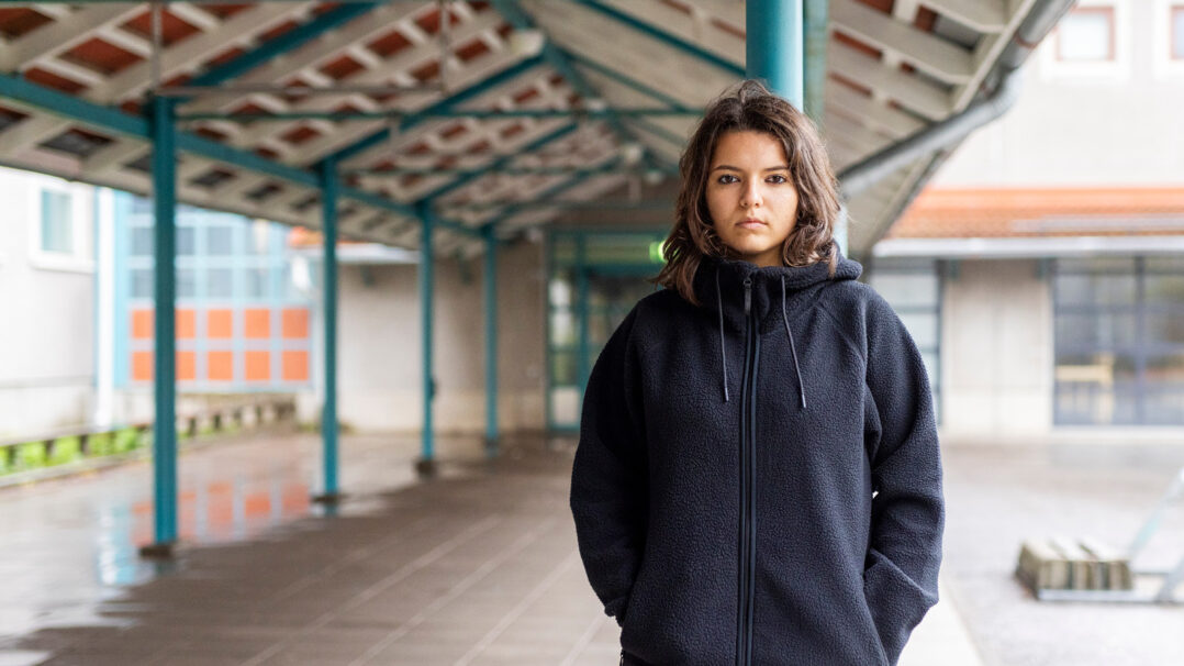 Tyttö katsoo suoraan kameraan vakavalla ilmeellä. Taustalla käytävä.