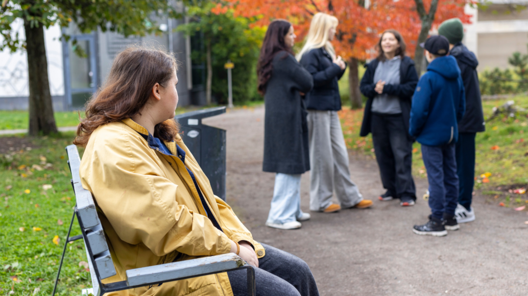 Puistomaisessa ympäristössä penkillä istuva henkilö katsoo kohti neljän hengen ryhmää, joka seisoo hiekkapolulla. Taustalla näkyy vihreää nurmikkoa, puita, joista osa on syksyn väreissä, sekä rakennuksen seinä.