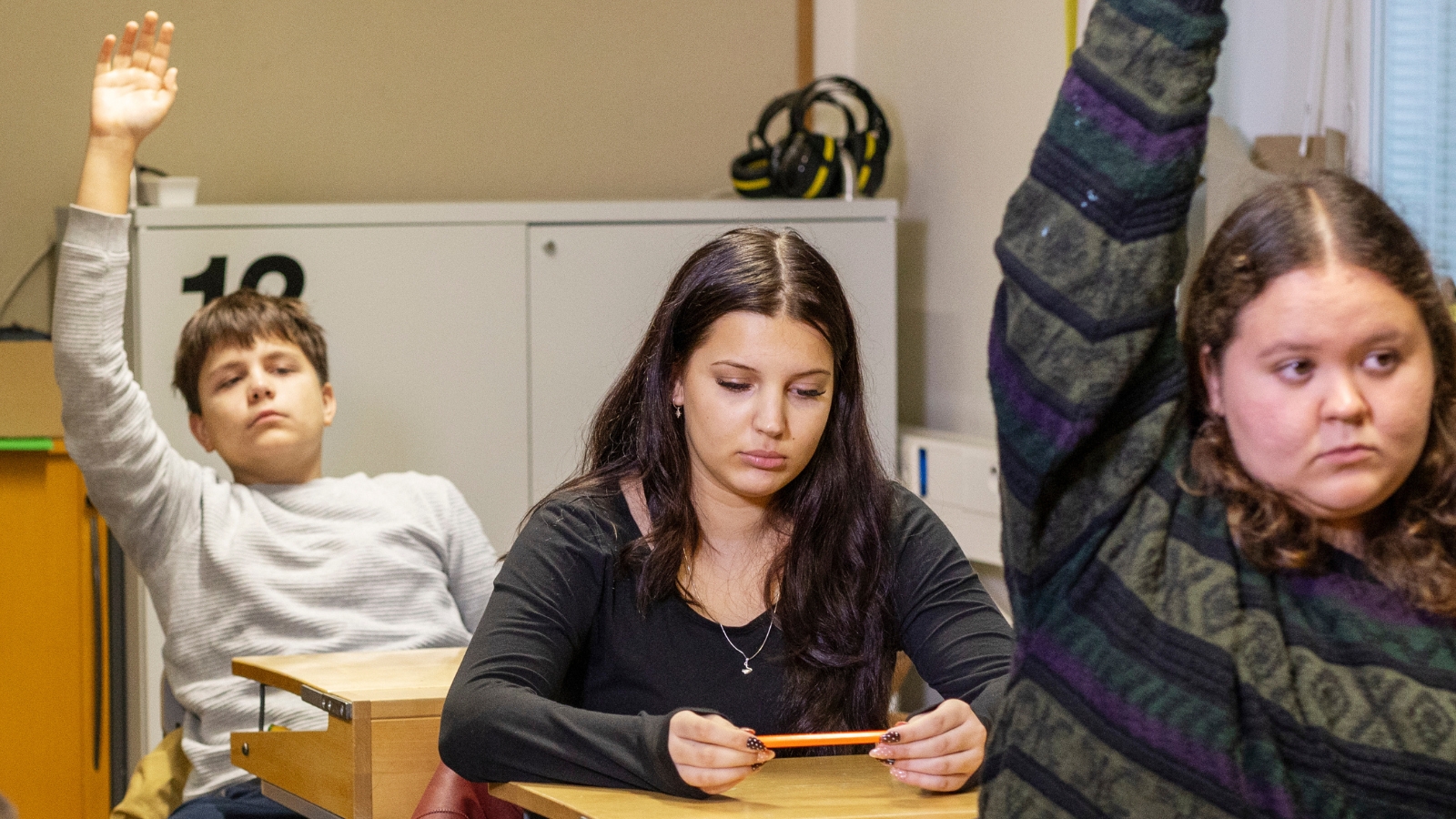 Kuvituskuva Lapsen ääni -kyselyn tulosten viestinään varten. Kuvassa näkyy luokkahuoneessa istuvat kolme nuorta, joista keskimmäinen katsoo murheellisena alas ja kaksi reunimmaista viittaavat.